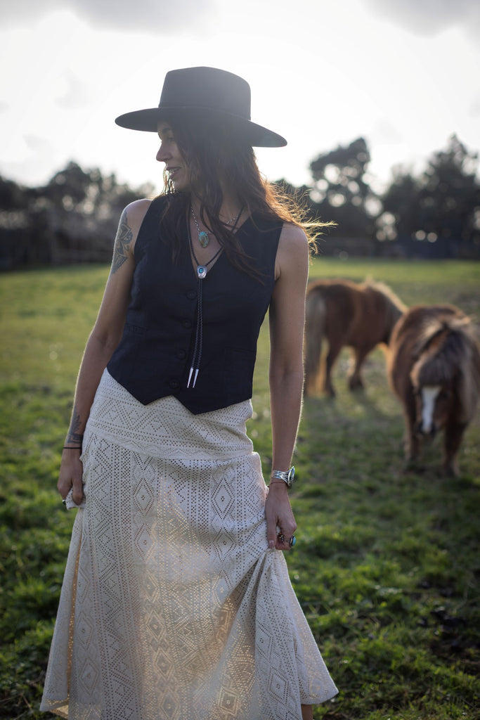 Woman in a black hat and vest standing in a field with horses in the background