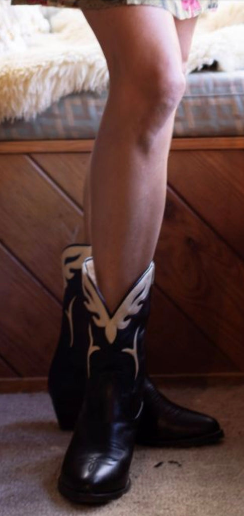 Person wearing black cowboy boots with white designs on a carpeted floor.