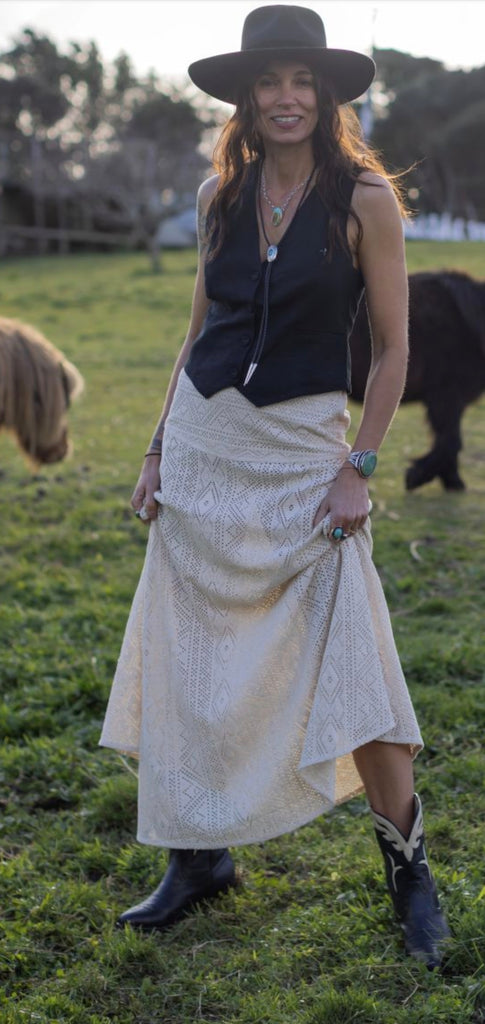 Woman in a black top and beige skirt  and cowboy boots standing in a field with animals in the background