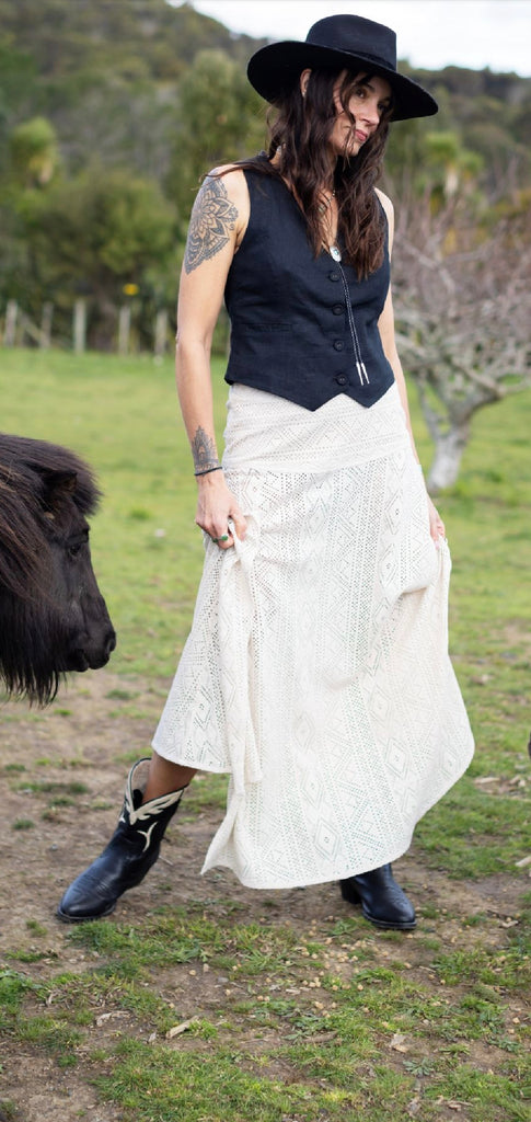 Woman in a black top and white skirt standing in a grassy field with a small black animal nearby.